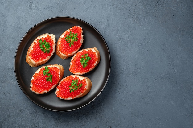 Panini con caviale rosso in un piatto nero su uno sfondo blu scuro. Antipasto di mare.