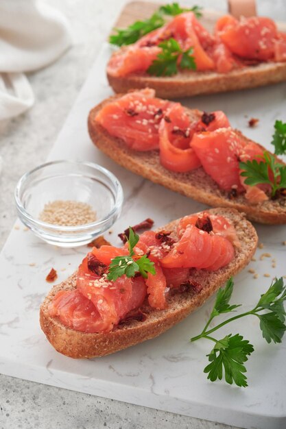 Panini con salmone salato Panini aperti con cereali o pane di segale integrale con salmone salato semi di sesamo e pomodoro secco su supporto in marmo bianco Frutti di mare Cibo sano Cucina scandinava