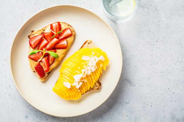 Panini della frutta del vegano con mango, fragola e burro di arachidi sul piatto bianco, vista superiore.