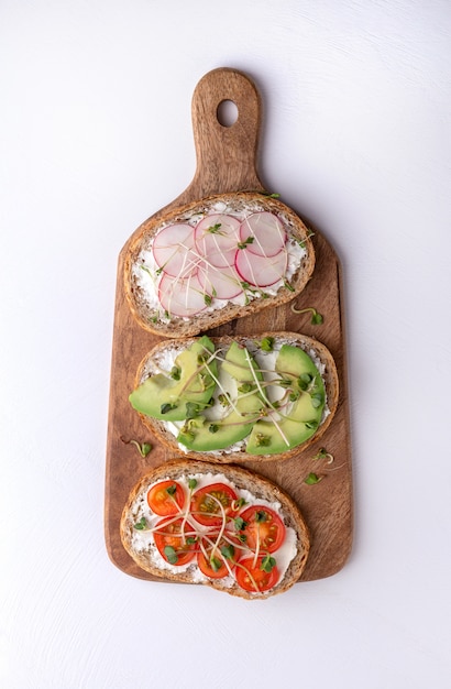 Panini di cereali con microgreens, ravanelli, avocado e pomodori su un tagliere su una superficie bianca