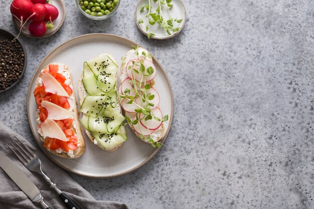 Panini su pane tostato con ingredienti, verdure, ravanelli, pomodori, cetrioli e microgreens su grigio. Vista dall'alto