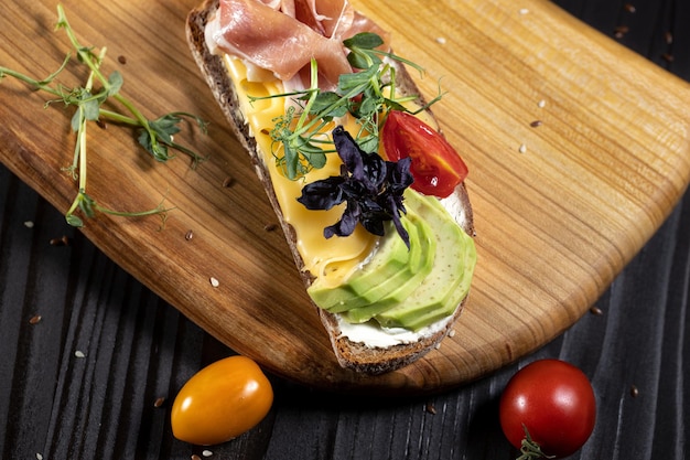 Panino aperto gustoso con prosciutto di avocado e pomodoro ciliegino su un piatto di legno