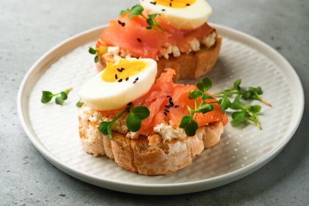 Panino con condimenti deliziosi salmone affumicato uova erbe e microverdi ravanello semi di sesamo neri su piastra bianca su sfondo grigio tavolo in cemento Superfood sano sandwich aperto Vista dall'alto