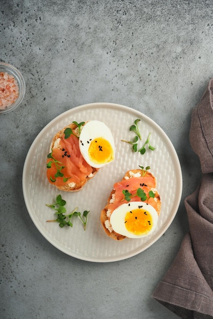 Panino con condimenti deliziosi salmone affumicato uova erbe e microverdi ravanello semi di sesamo neri su piastra bianca su sfondo grigio tavolo in cemento Superfood sano sandwich aperto Vista dall'alto