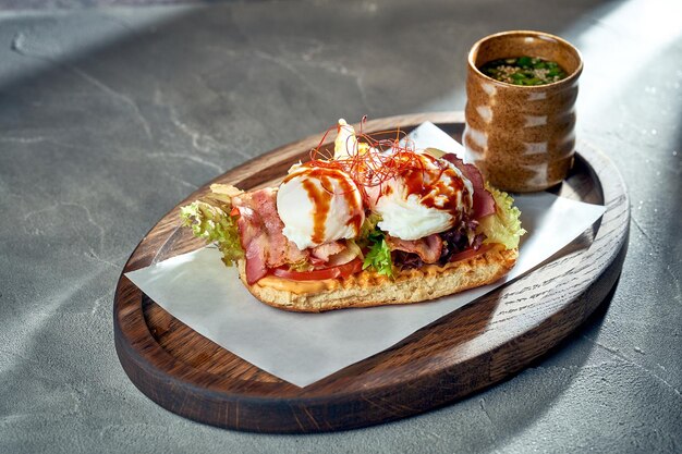 Panino con pancetta di pomodoro e uovo in camicia su pane bianco Sfondo concreto Uova Benedict per colazione Luce del mattino con ombre dure