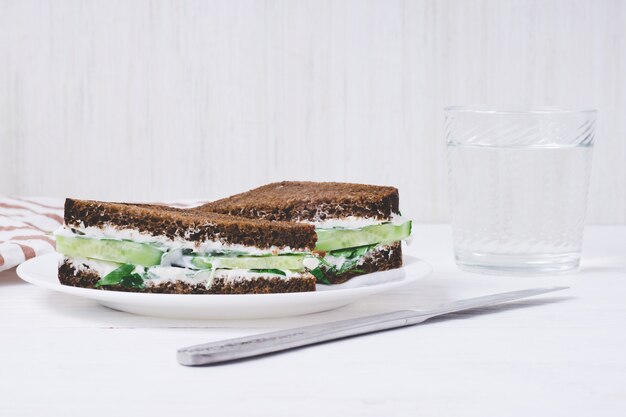 Panino con pane nero, cetriolo e crema di formaggio su un piatto su sfondo bianco.