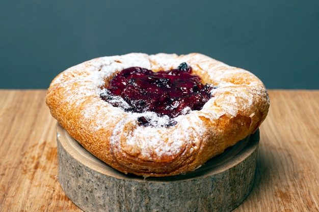 Panino della pasta sfoglia con i cherrys e la crema pasticcera spruzzati con zucchero in polvere su un supporto di legno della foresta su un primo piano grigio del fondo.