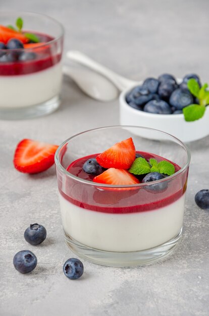 Panna cotta dolce italiano in un bicchiere con salsa di frutti di bosco, fragole fresche e mirtilli in cima su uno sfondo grigio cemento.