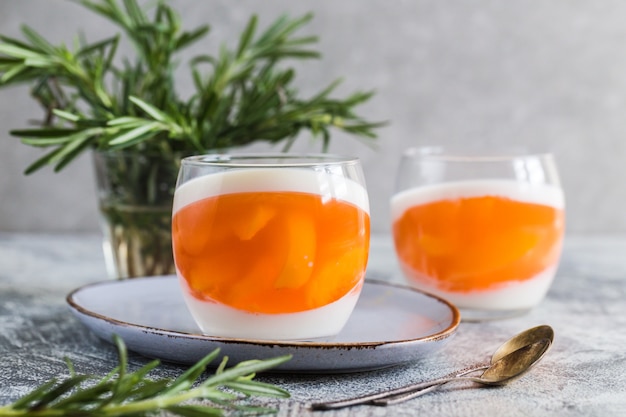 Panna cotta fatta in casa con fette di pesca e gelatina di pesche in vasetti di vetro su fondo grigio cemento.