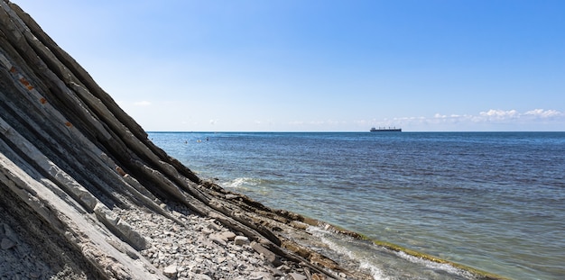 Panorama del paesaggio marino. Pittoresca spiaggia di pietra selvaggia ai piedi delle scogliere, cielo azzurro con nuvole e una nave all'orizzonte. Dintorni della località di Gelendzhik. Russia, Mar Nero