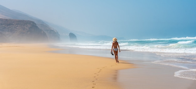 Panoramica di un giovane turista con un cappello che cammina da solo sulla spiaggia di Cofete del parco naturale di Jandia, Barlovento, a sud di Fuerteventura, Isole Canarie. Spagna