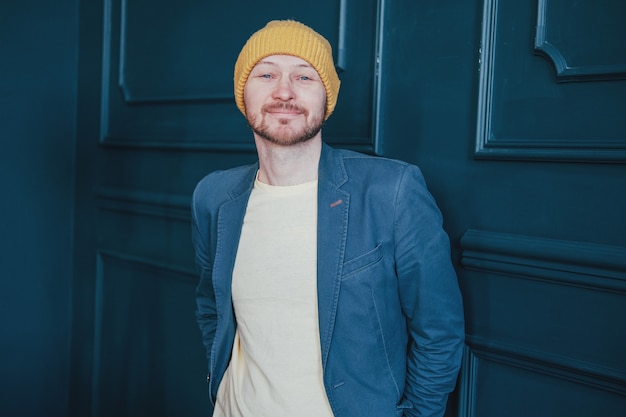 Pantaloni a vita bassa barbuti adulti attraenti dell&#39;uomo in cappello giallo che sembra amichevole alla macchina fotografica sulla parete blu