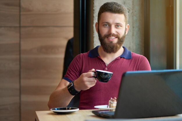 Pantaloni a vita bassa sorridenti che tengono la tazza di caffè, funzionante con il computer portatile in caffè.