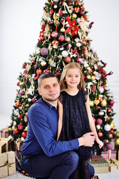 Papà abbraccia la sua piccola figlia in un abito elegante sullo sfondo di un albero di Natale.