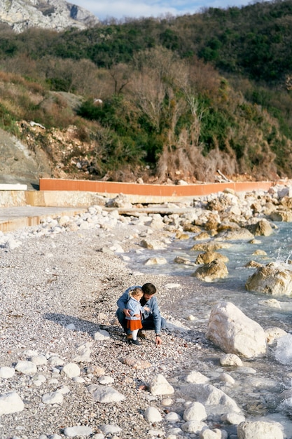 Papà accovacciato accanto a una bambina raccoglieva sassi da una spiaggia rocciosa vicino all'acqua