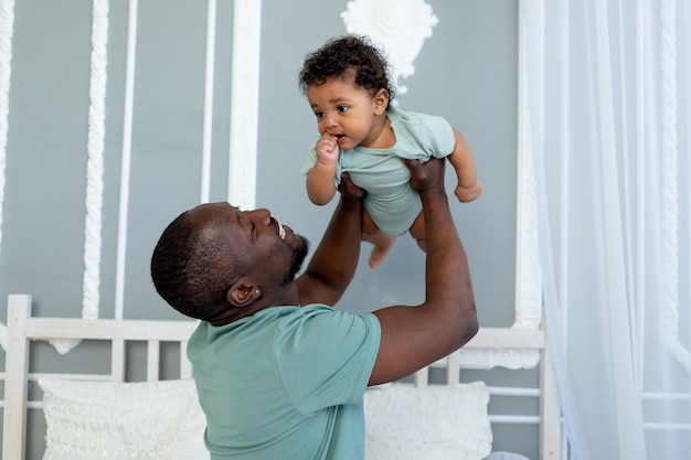 Papà afroamericano sorridente felice con il figlio del bambino sul letto a casa che stringe a sé e che gioca, famiglia felice