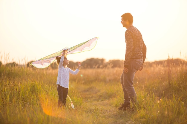 Papà aiuta sua figlia a far volare un aquilone in un campo in estate al tramonto. Animazione per famiglie all'aperto, festa del papà, festa dei bambini. Aree rurali, sostegno, assistenza reciproca. Luce arancione del sole