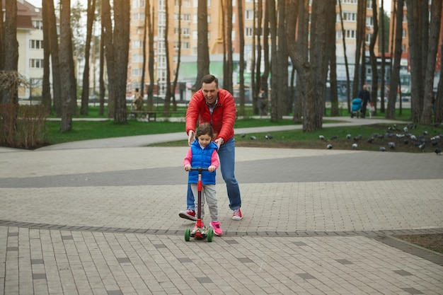 Papà allegro che corre accanto alla sua figlia carina in sella a uno scooter a spinta godendosi una giornata libera in un parco cittadino Festa del papà Concetto di infanzia felice