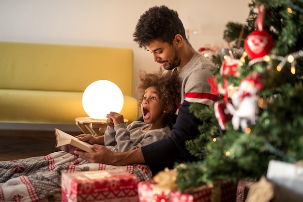 Papà che legge le fiabe a sua figlia sdraiato accanto all'albero di Natale