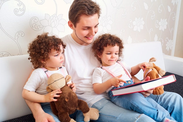 Papà con due figlie gemelle che leggono un libro sul divano in camera insegnando ai bambini a leggere una famiglia felice