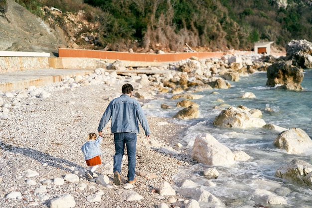 Papà con una giacca di jeans e jeans cammina con una bambina che le tiene la mano lungo una spiaggia di ciottoli rocciosa