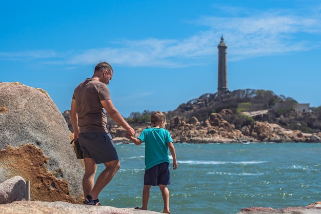 Papà cura Figlio vicino al mare passeggiata trascorrere del tempo insieme esplorare nuovo Uomo tenere bambino mano sfondo faro Simbolo della coscienza individuale sensibilizzazione ragazzo padre famiglia unione influenza nel bambino felice dal vivo