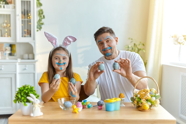 Papà e figlia facce macchiate di vernice blu per dipingere le uova. sul tavolo c'è un cesto con uova di Pasqua e colori.