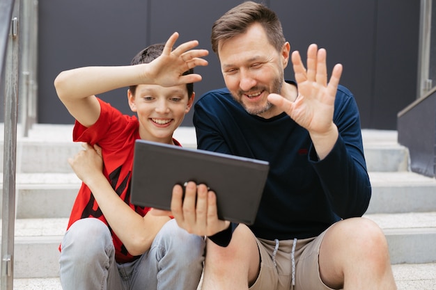 Papà e figlio fanno una videochiamata usando il tablet in città su una scala di cemento, agitano allegramente le mani dicendo ciao