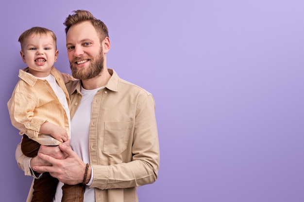 Papà felice e piccolo figlio che posano alla macchina fotografica sorridente isolato sulla parete viola