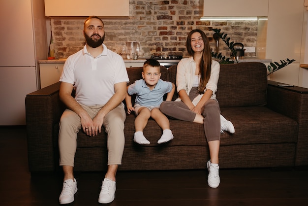 Papà, figlio e mamma stanno guardando la TV sul divano dell'appartamento. La famiglia si sta godendo la serata. Parenti in casa. Una giovane madre con i capelli lunghi sta ridendo.