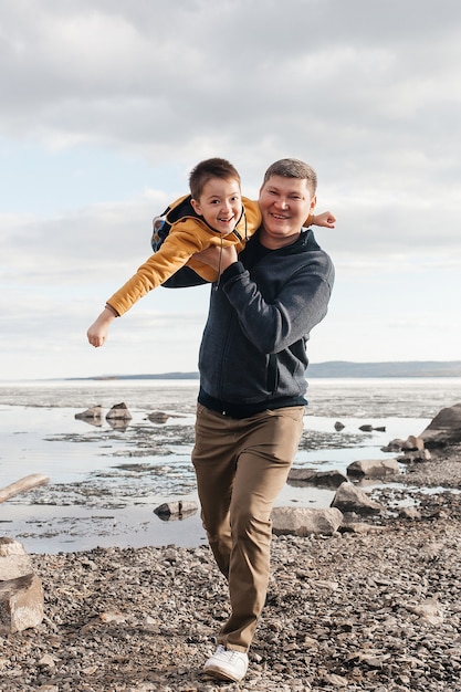 Papà gioca con suo figlio sulla riva del fiume. Un bellissimo padre e figlio si divertono a giocare e sorridere in una giornata di sole. Il bambino è tra le braccia di suo padre. Ricreazione all'aperto.