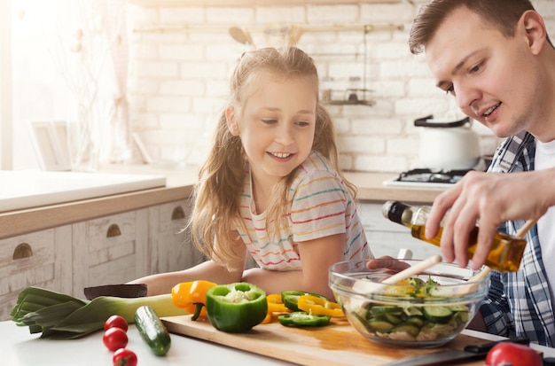 Papà positivo e figlia piccola che cucinano in cucina a casa. Uomo che insegna alla ragazza ad aggiungere il condimento per l'insalata, copia spazio