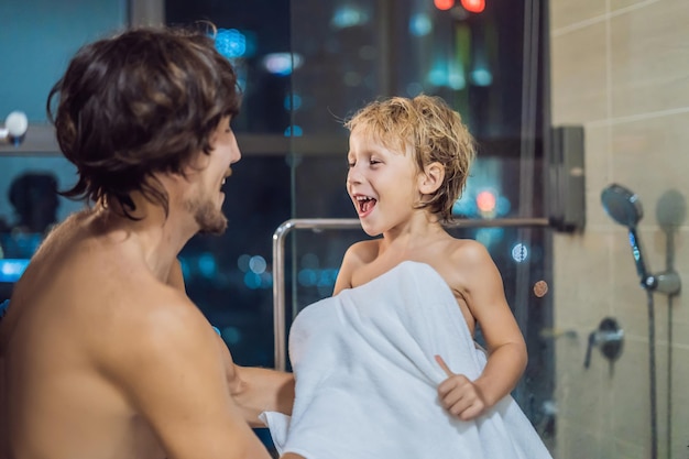 Papà pulisce il figlio con un asciugamano dopo la doccia la sera prima di andare a dormire sullo sfondo di una finestra con vista panoramica sulla città