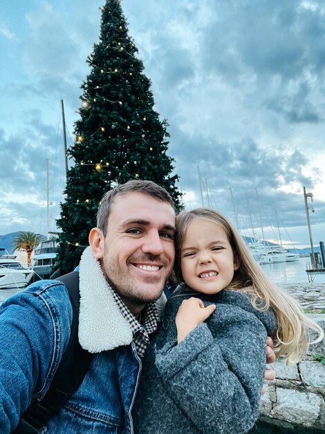 Papà sorridente con la bambina vicino all'albero di Natale decorato sul molo