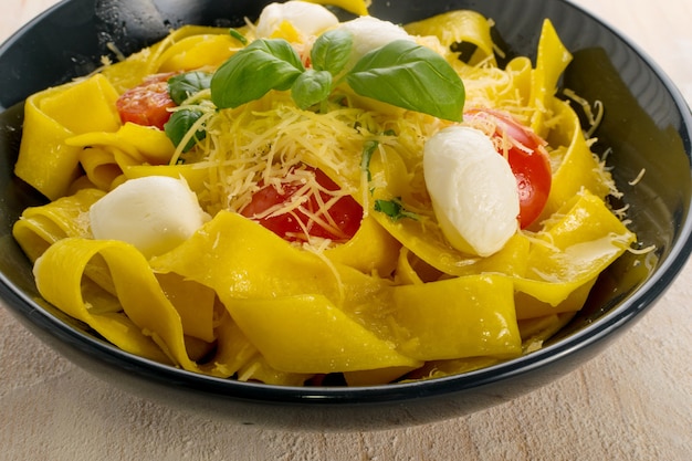 pappardelle di pasta gialla cotta, fettuccine o tagliatelle in ciotola nera. Tagliatelle fatte in casa all'uovo o maccheroni con pomodoro, basilico e palline di mozzarella