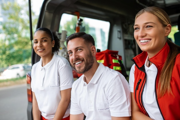 Paramedici e medico seduti sul retro dell'ambulanza Gruppo di tre paramedici in piedi davanti all'ambulanza con un sorriso