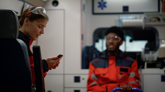 Paramedico sorridente che parla con un medico afro in un'auto di emergenza Medici di emergenza che guidano una chiamata al 911 Assistente di ambulanza femminile che scrive un messaggio sul telefono cellulare Concetto medico