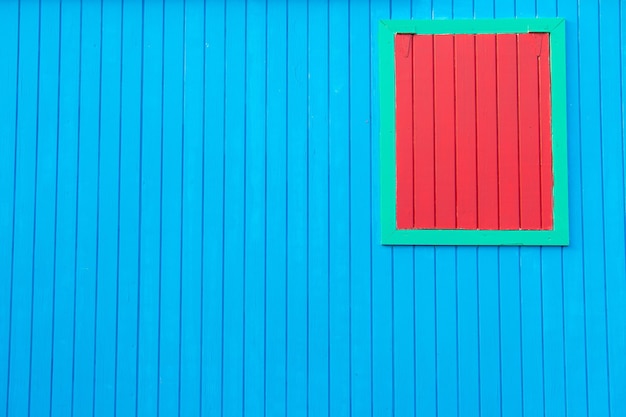 Parete blu di legno variopinta luminosa della casa con la finestra rossa chiusa nel telaio verde come fondo astratto strutturato, spazio della copia