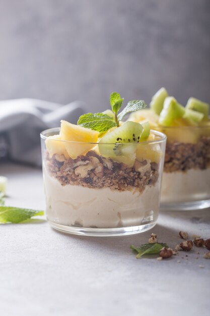 Parfait di yogurt fatto in casa con muesli, kiwi, ananas e noci in un bicchiere per una sana colazione su sfondo concreto