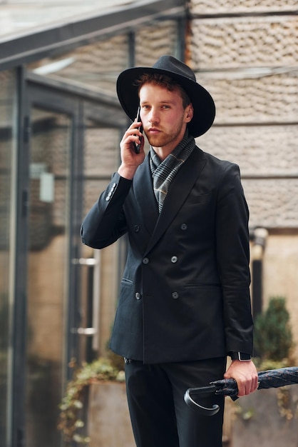 Parlando al telefono Elegante giovane con cappello nero e vestiti con ombrello in mano all'aperto in città