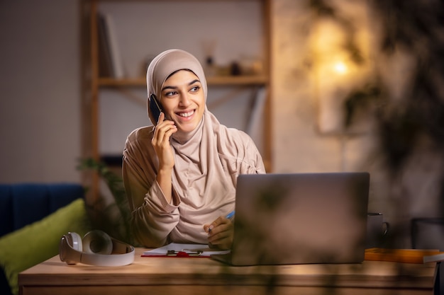 Parlando al telefono. Felice donna musulmana a casa durante la lezione online. Tecnologie moderne, educazione remota, etnia e concetto di tradizione. Emozioni umane, stile di vita. Utilizzando laptop seduto a casa.