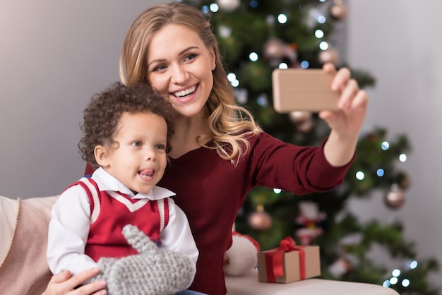 Parlando di selfie. Bella donna abbastanza gioiosa seduta sul divano con suo figlio e tenendo il suo smartphone mentre prende selfie