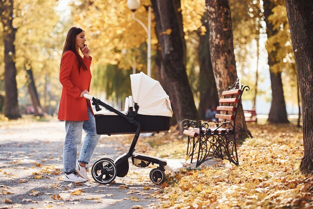 Parlare al telefono. La madre in cappotto rosso fa una passeggiata con il suo bambino nella carrozzina nel parco in autunno.