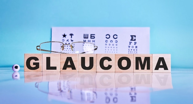 Parola GLAUCOMA scritta su cubi di legno, occhiali, occhi sullo sfondo di un tavolo di prova dell'occhio.