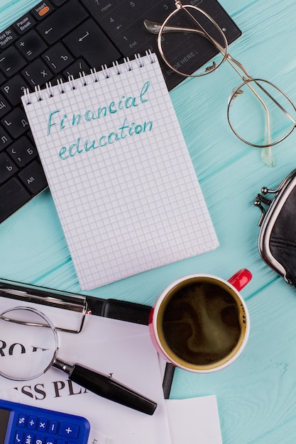 Parole di educazione finanziaria su blocco note, tastiera e tazza di caffè sulla scrivania blu chiaro. Vari articoli per ufficio sono costituiti da appunti con lente d'ingrandimento calcolatrice.