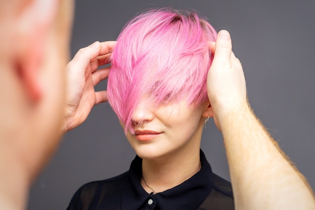 Parrucchiere che controlla breve acconciatura rosa di giovane donna sul muro grigio