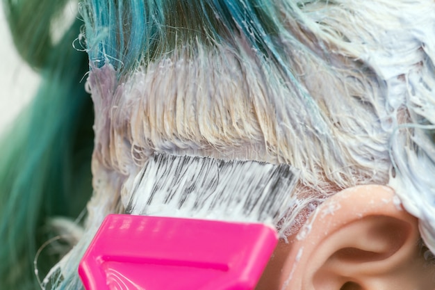 Parrucchiere che usa la spazzola rosa mentre applica la vernice alla cliente femminile con il colore dei capelli color smeraldo durante il processo di sbiancamento delle radici dei capelli