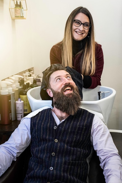 Parrucchiere donna che asciuga i capelli del cliente hipster dopo il risciacquo al lavabo Uomo sorridente hipster al salone del barbiere che si fa tagliare barba e capelli Concetto del negozio di barbiere