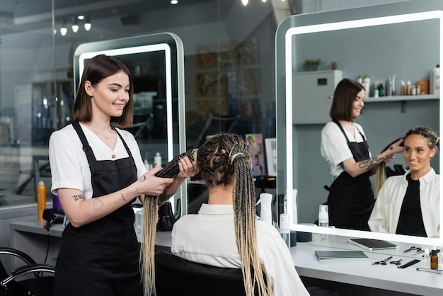 parrucchiere e cliente salone di bellezza stilista di capelli tatuati che fa i capelli di una donna allegra con trecce