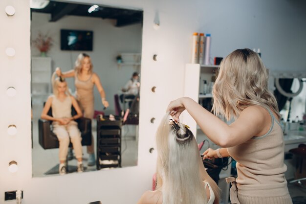 Parrucchiere femminile che fa le estensioni dei capelli alla giovane donna con capelli biondi nel salone di bellezza. Extension professionale per capelli.